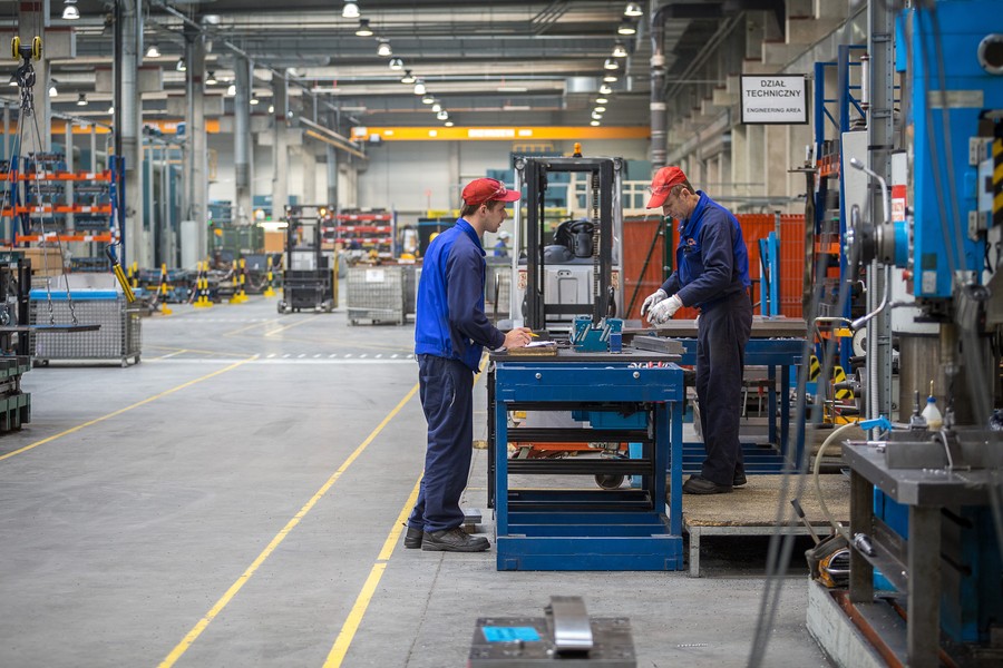 Dwóch panów pracujących przy stole narzędziowym na hali zakładowej ubrani w niebieskie stroje robocze i czerwone czapki, fot. Szymon Zdziebło www.tarantroga.pl dla UMWKP