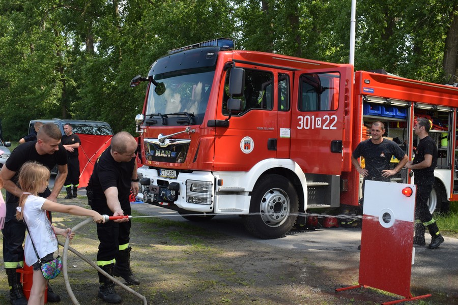 Na pierwszym planie mała dziewczynka w towarzystwie strażaków polewa wodę z węża w kierunku specjalnej tarczy do ćwiczeń z czerwonym sygnalizatorem. Na drugim planie duży czerwony wóz bojowy Straży Pożarnej.