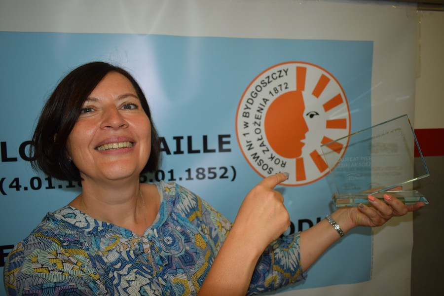 Laureatka konkursu ENEA Akademia Talentów 2018; Foto Jacek Knychała, Na tle planszy zawierającej logo bydgoskiego Ośrodka im. L. Braille’a stoi nauczycielka, która w lewej ręce trzyma statuetkę konkursu Enea Akademia Talentów 2018 (Kategoria: Sztuka)