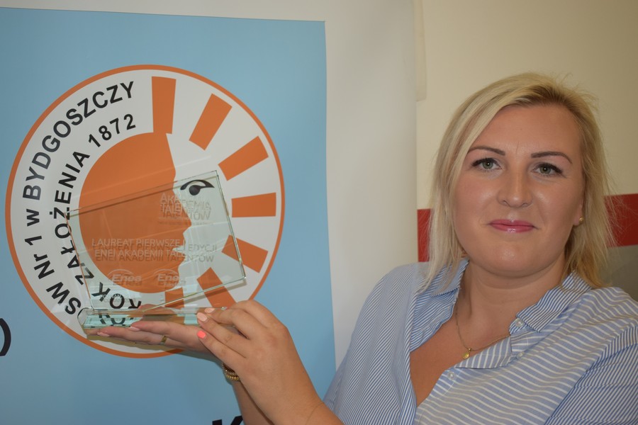 Laureatka konkursu ENEA Akademia Talentów 2018; Foto Jacek Knychała, Na tle planszy zawierającej logo bydgoskiego Ośrodka im. L. Braille’a stoi nauczycielka, która w prawej ręce trzyma statuetkę konkursu Enea Akademia Talentów 2018 (Kategoria: Sport)