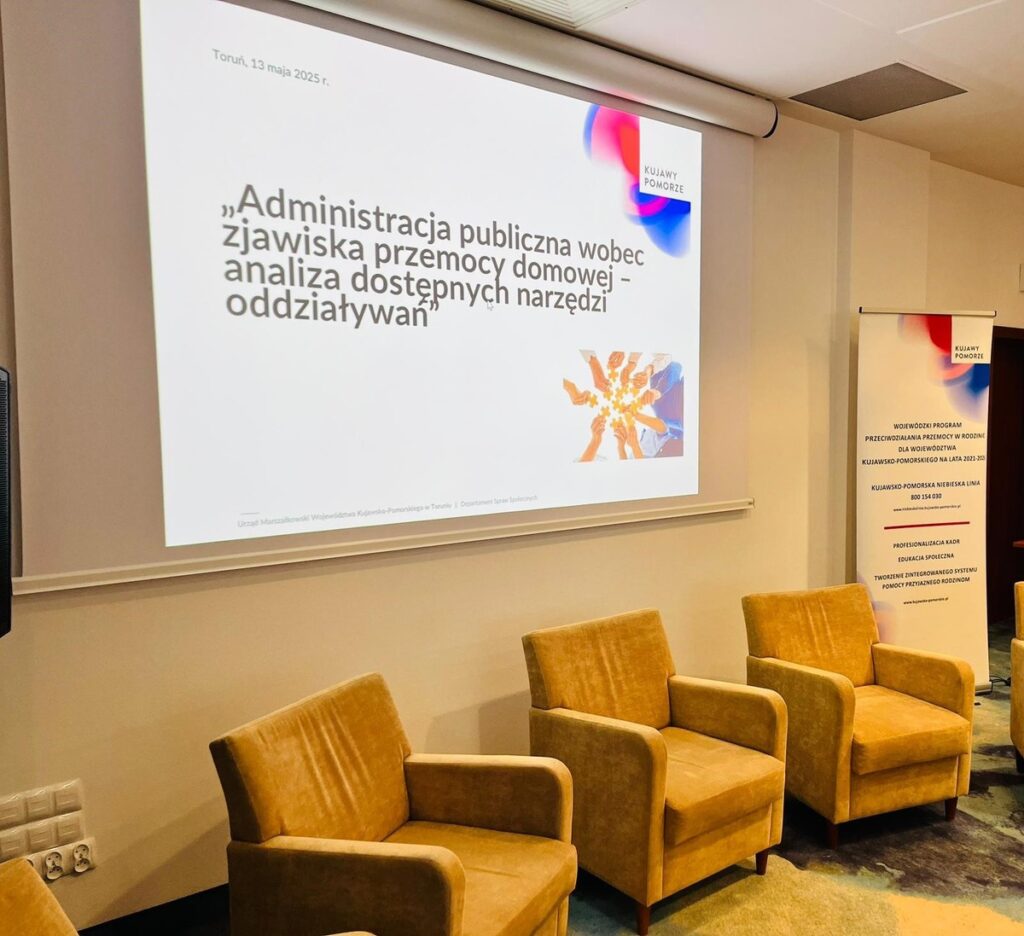 Tablica projekcyjna z tytułem seminarium „„Administracja publiczna wobec zjawiska przemocy domowej – analiza dostępnych narzędzi oddziaływań”, fotele ustawione przy ścianie, roll-up Kujawsko-Pomorskiej Niebieskiej Linii, fot. Biuro Wsparcia Rodziny, Przeciwdziałania Przemocy i Uzależnieniom