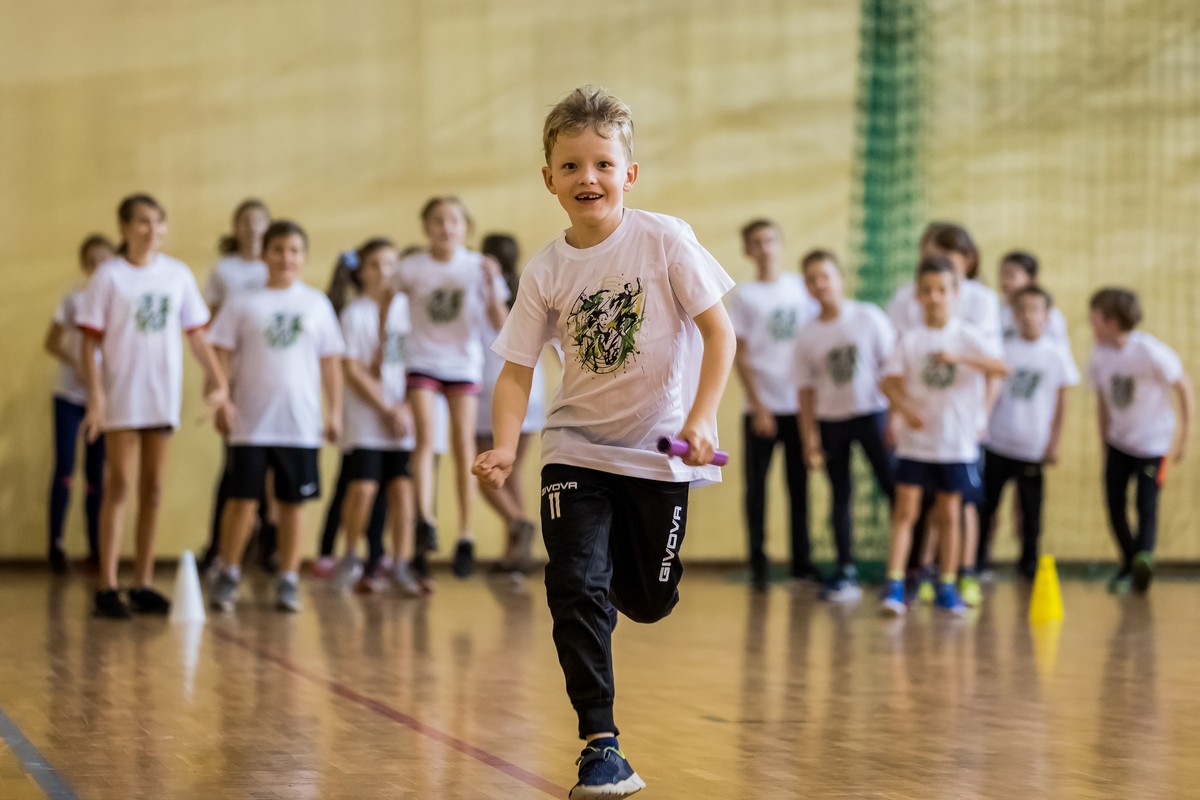 Biegnący chłopiec podczas sztafety na hali sportowej, w tle inne dzieci kibicujące.